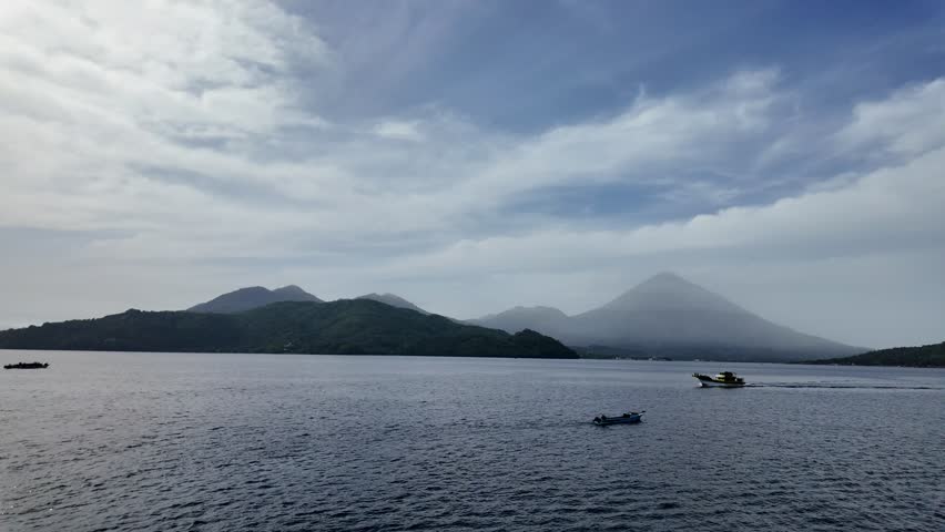 4K 60fps wide landscape shot of a calm ocean bay with majestic misty volcanic mountains in the background and small local boats crossing the tranquil waters under a cloudy sky.