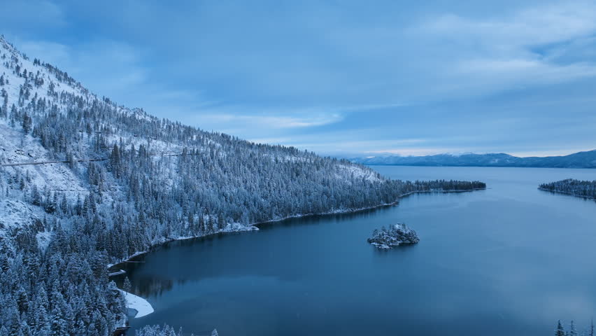 Top down drone view of Lake Tahoe during winter season. Snow covered forest lines the shoreline while deep blue water fills the frame, creating strong contrast and clean geometric natural composition.