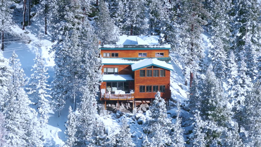 Cozy wooden cabin surrounded by snow covered pine forest during gentle fluffy snowfall. Winter mountain atmosphere with fresh powder and peaceful alpine mood. California, USA.