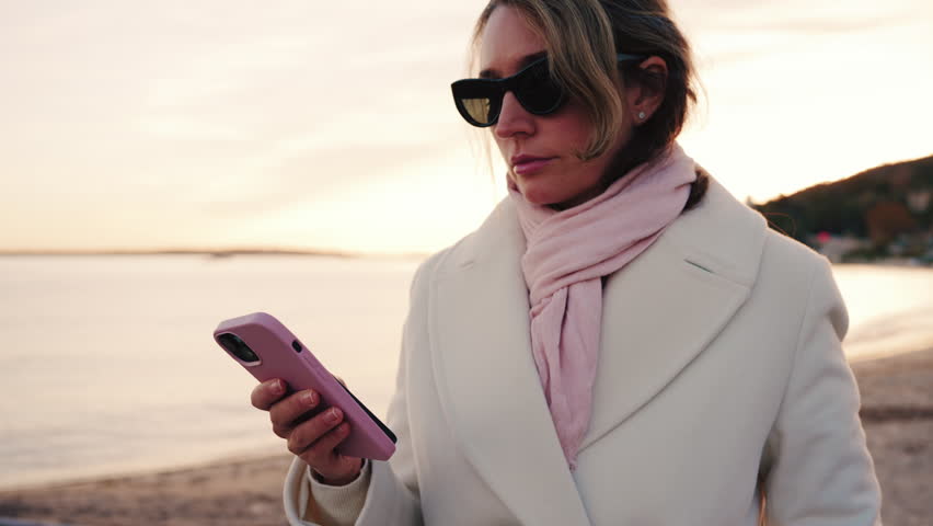 Elegant woman wearing a white coat, scarf, and sunglasses checks her phone while standing on a beach at sunset