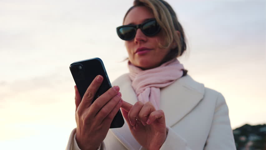 Elegant woman wearing a white coat, scarf, and sunglasses checks her phone while standing on a beach at sunset