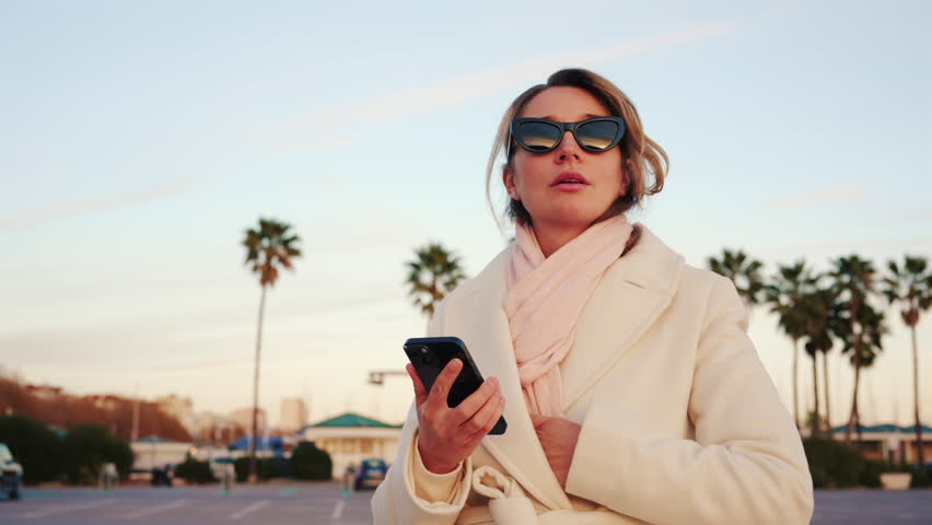 Elegant woman wearing a white coat, scarf, and sunglasses checks her phone in a sunny outdoor area lined with palm trees