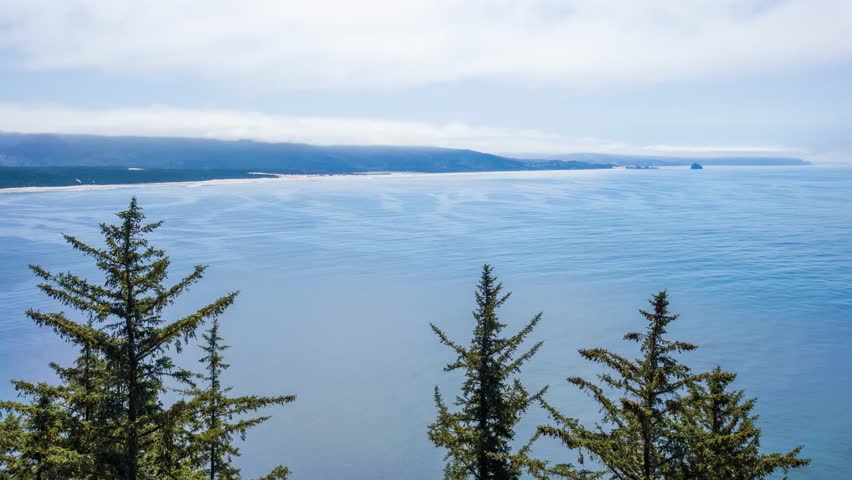 Scenic footage captures a high-angle, wide view of the rugged coastline at Cape Lookout State Park in Tillamook, Oregon. Lush green Sitka spruce trees frame the foreground, overlooking the vast, turqu