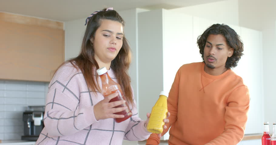 Diverse friends at counter debating ketchup or mustard until third friend shaking ketchup bottle. Collaboration, communication, casual, vibrant, social, domestic, interaction