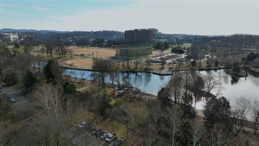Parthenon in Nashville, Tennessee surrounded by Centennial Park lake and walking paths. Iconic landmark with city skyline in background under clear daylight. Aerial approaching shot.