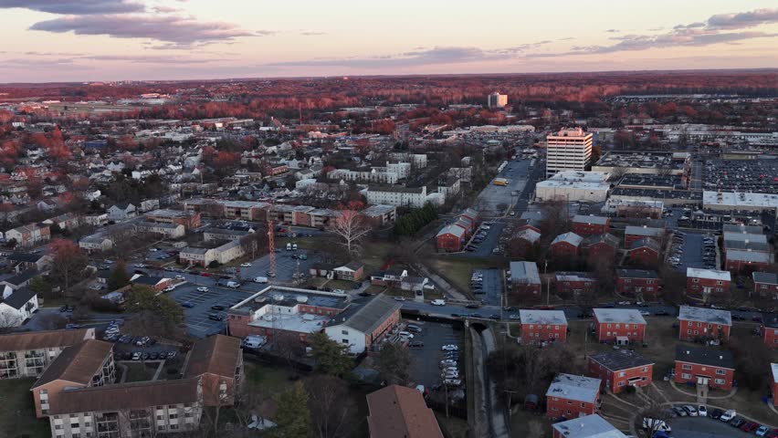 Aerial sunset flyover of Laurel, Maryland, showing residential neighborhoods, apartment complexes and commercial buildings glowing in warm winter light under calm evening sky. Wide shot.