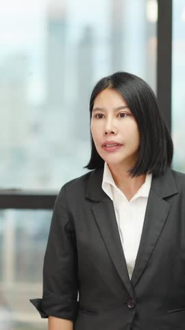 A cheerful Asian businesswoman in a blazer waves enthusiastically against a bright office window.