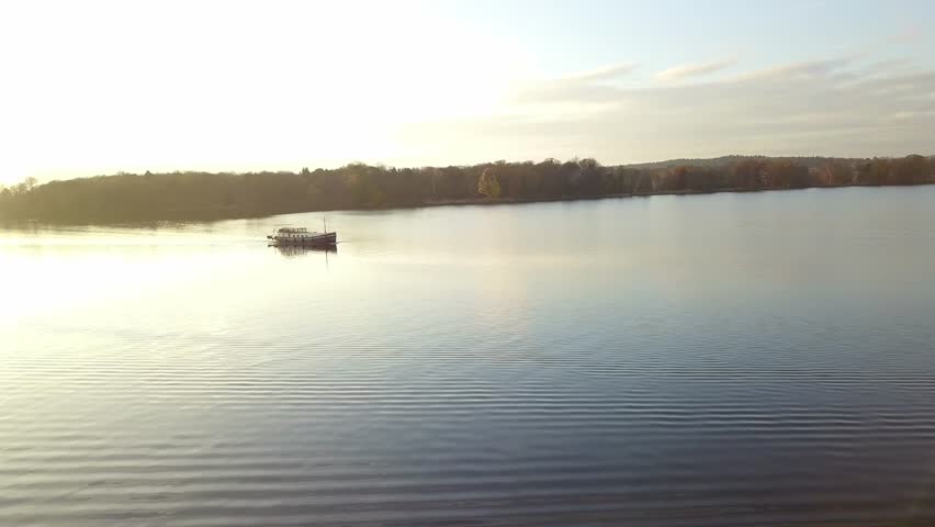 Aerial pan right of passenger boat navigating open lake under soft sunset sky with warm reflections on water surface.