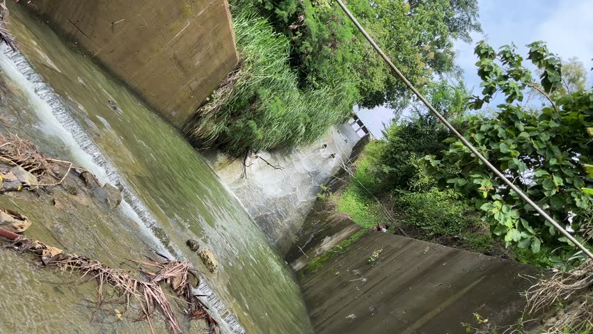 Wide view of water flowing through dam spillway canal with green vegetation on both sides. Urban water management infrastructure during rainy season representing flood control and environmental engineering.