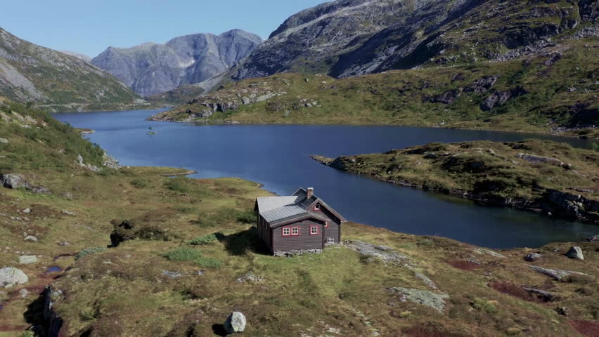 A serene view of a mountain landscape featuring a calm lake and a small cabin nestled among the hills. The scene captures the beauty of nature in a peaceful setting.