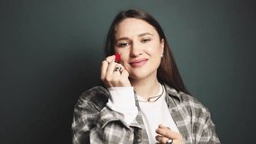 Smiling brunette woman applying bright blush stick on cheekbones and nose, blending with finger, natural makeup routine in motion, beauty cosmetic product demonstration on gray green background studio - Powered by Shutterstock - Get 15% off with code: PIKWIZARD15