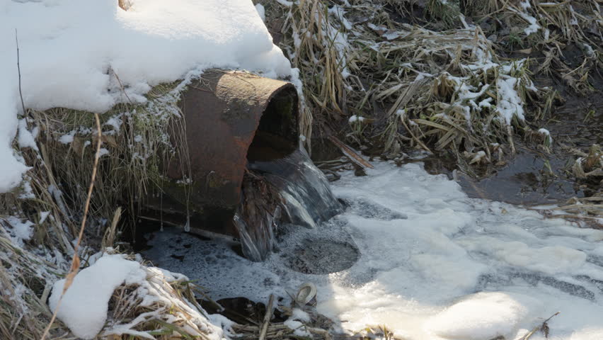 White foam accumulates on water surface near metal drainage pipe discharge. Bubbles floating in the stream during winter thaw. Environmental view with audio.