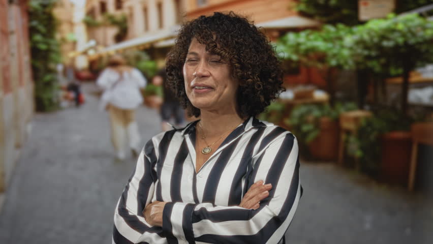Hispanic woman in black and white striped blouse crosses arms on cobblestone street lined with potted greenery at outdoor cafe; rejection.