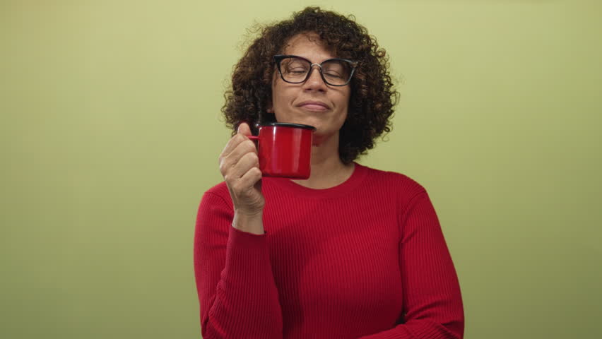 Woman holding red mug near chin with glasses and curly hair in studio with light green background; calm reflection.