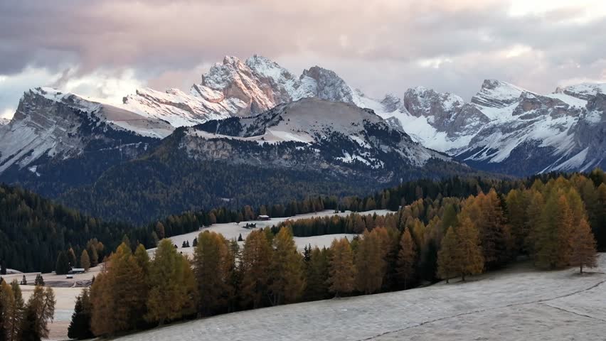 Stunning drone view of the Sassolungo mountain range, Langkofel in the Dolomites. Majestic alpine peaks covered in light snow, autumn colored forest all around, and yellow grass covered in morning white hoarfrost. Picturesque natural landscape