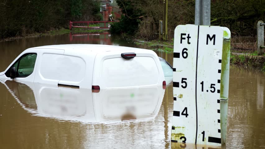 Flood warning sign measuring water level while van remaining submerged on rural road. Brown river water rising and surpassing gauge markings during severe storm. Climate crisis worsening and causing