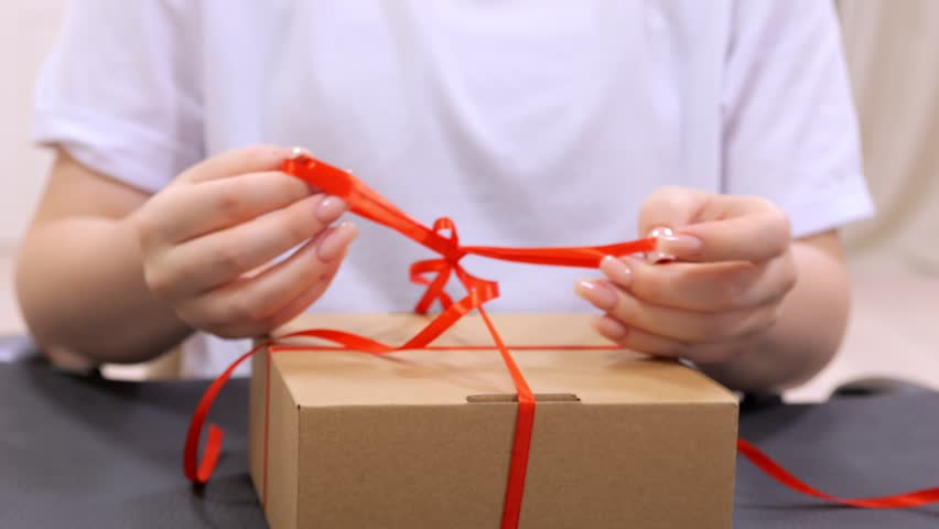 Person is tying a red ribbon around a brown box. The ribbon is tied in a bow