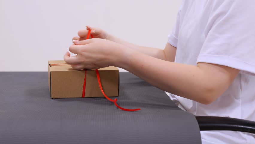 Person is tying a red ribbon around a box. The box is brown and is sitting on a table