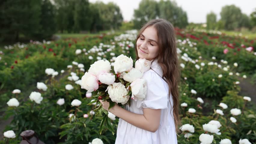 portrait beautiful young girl in white dress with flowers in a peony field in summer day. Smiling happy girl holding flowers pink peonies bouquet in a blooming spring garden. Copy space.