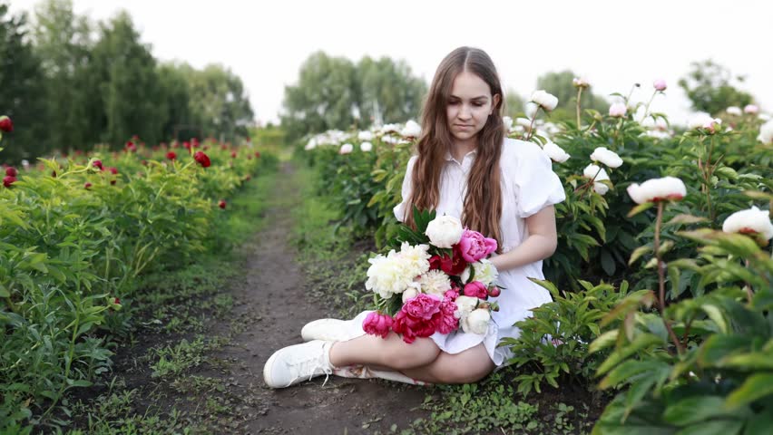 beautiful young girl in white dress sit with flowers in a peony field in summer day. Smiling happy girl holding flowers pink peonies bouquet in a blooming spring garden. Copy space.