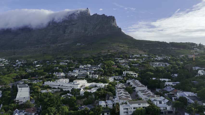 Drone flies south toward Table Mountain as clouds pour over the top at sunset in Cape Town, South Africa