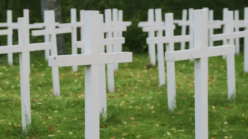 Numerous white crosses are planted in straight rows in a grassy field with fallen leaves. Memorial or grave markers for military or civilian casualties in a Cemetery