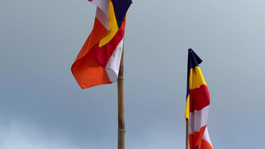 Buddhist flag moves on a high flagpole with a gray clouded sky behind, showing steady wind conditions above a peaceful outdoor setting. Religious banner.