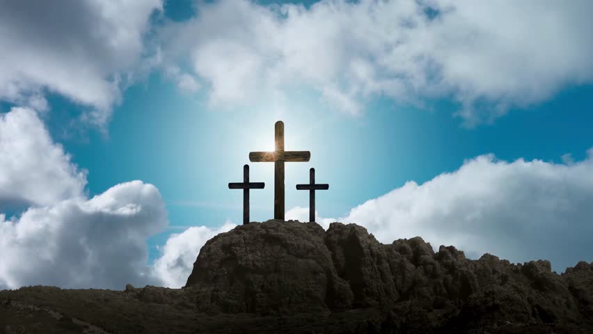 Three crosses stand atop a rocky hill against a vibrant blue sky filled with puffy white clouds. Sunlight shines through the central cross, creating a symbolic and spiritual atmosphere.