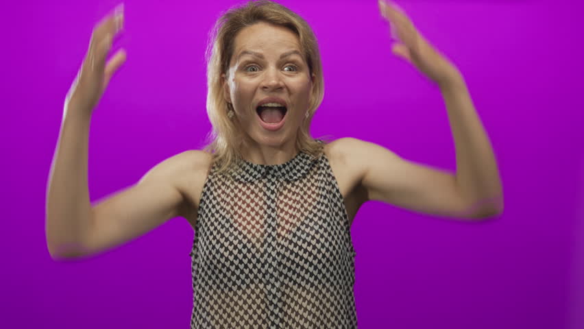 Woman clenches fists and smiles with closed eyes in vibrant purple studio setting wearing patterned sleeveless top; triumph victory celebration joy.
