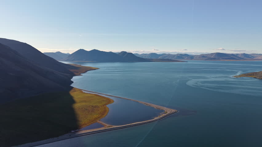 Aerial view side tracking follows curved lagoon and causeway at Rumilet Bay, Chukotka. Morning light highlights tundra colors and still water. Remote arctic landscape for exploration and travel