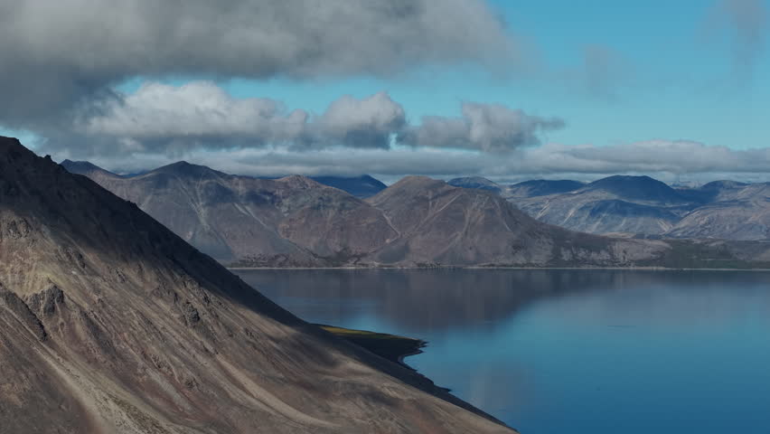 Slow drift reveals rocky ridges above Rumilet Bay in Chukotka. Dramatic clouds hang over distant peaks and calm water. Early autumn light adds atmosphere for nature and tourism projects