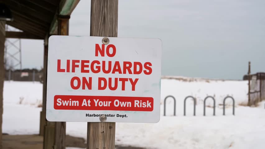 Winter beach covered with snow with No Lifeguard On Duty, Swim At Your Own Risk sign flapping in the wind