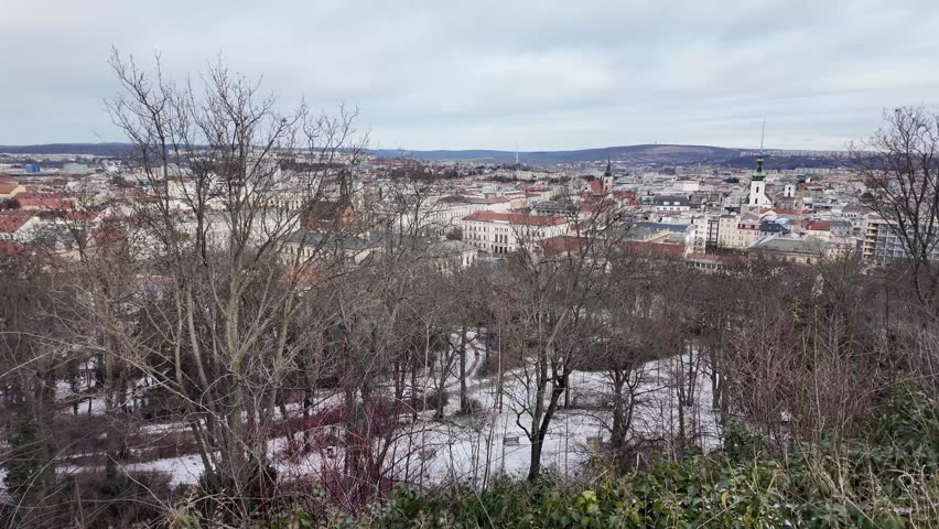 Cinematic slow motion panning shot of Brno city center covered in snow, viewed from the Spilberk fortress. The footage highlights the red rooftops dusted with white, the iconic cathedral spires, and the soft winter light over Moravia.