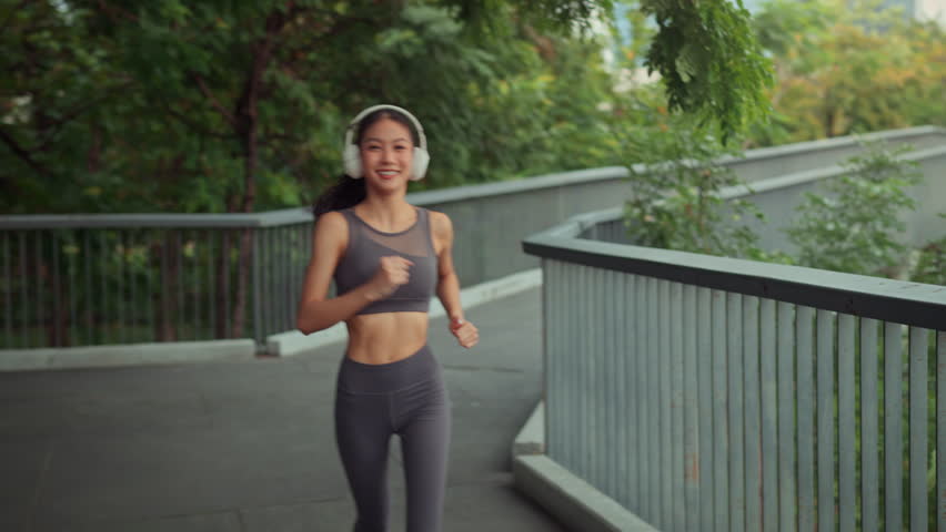 Smiling beautiful Asian woman running in city park wearing wireless headphones, happy young lady jogging on forest bridge for cardio workout, concept of healthy lifestyle, urban fitness, morning exercise, and wellness in a green environment.