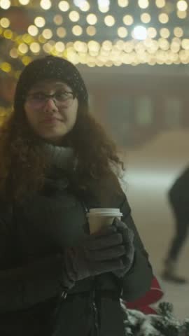 Female Individual Consumes Steaming Hot Coffee. Young Woman Experiencing Winter Evening At Lively Coffee Stall. Female Patron Enjoys Warm Cup Of Coffee During Winter Night Market Event