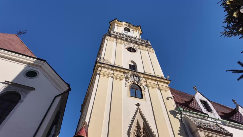 Cinematic slow motion shot looking up at the historic clock tower of the Old Town Hall in Bratislava, Slovakia. Highlights include the ornate architecture and the iconic colorful mosaic roof tiles.