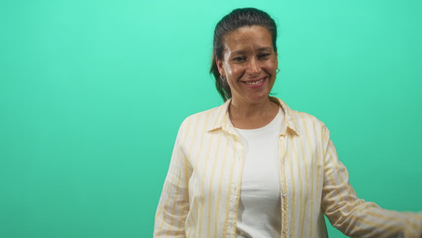 Woman points finger to nose in studio wearing yellow striped shirt and white tee, smiling with ponytail; playful joy.