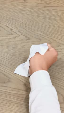 Close up in indoor lighting of Asian man with cerebral palsy holding glasses temple with left hand, wiping lens with white cloth in right hand on wooden table. Fine motor skills training.