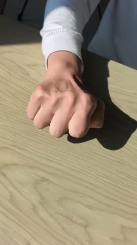 Front side view in indoor sunlight of Asian man with cerebral palsy extending and clenching right hand fist with index finger bent on wooden table. Fine motor skills training.