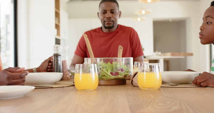 African American family clasping hands over salad at table after reaching across bowing in prayer. Family, bond, unity, nourishment, togetherness, warmth, domestic