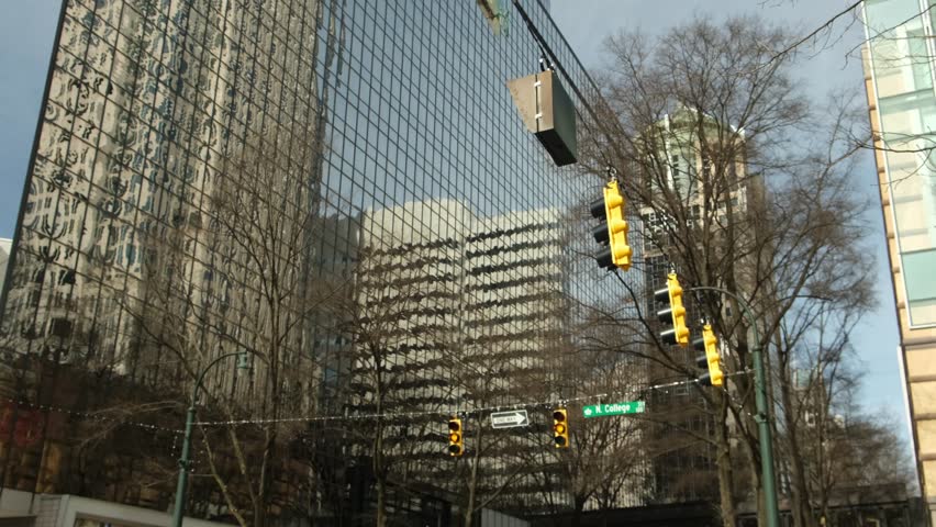 People walk along the street near modern buildings. Traffic lights change colors. The glass building reflects its surroundings. Cars drive by the busy street.