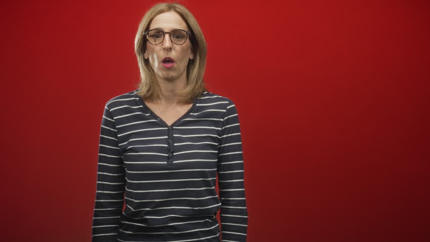 Woman covering mouth with hands in red studio wearing glasses and striped sweater; surprise reaction.