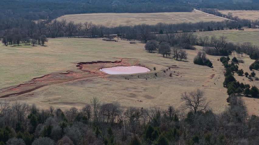 Aerial drone view of rural Oklahoma pasture with red dirt pond erosion, small water reservoir, and winter grassland landscape in open countryside.