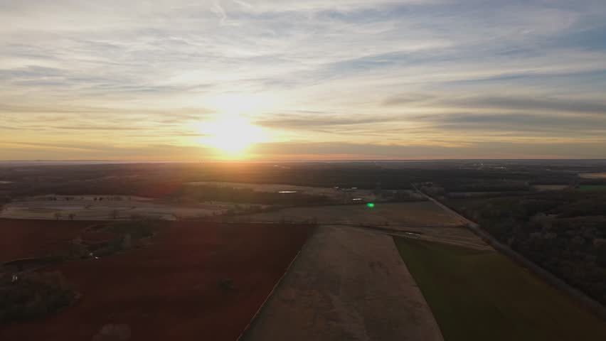Aerial drone view of Oklahoma farmland at sunset with patchwork agricultural fields and glowing horizon over rural countryside.