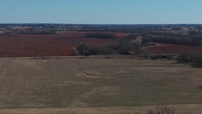 Aerial drone view of rural Oklahoma farmland with green crop field, red soil pasture, and small farm truck traveling along country road.
