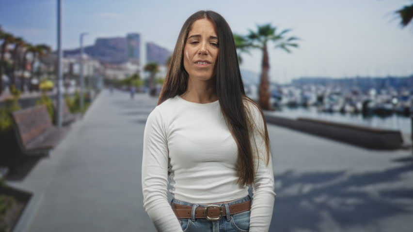 Woman smiling with long hair and hands at waist on a sunny street by a marina, wearing white shirt and jeans, facing camera; joyful confidence.