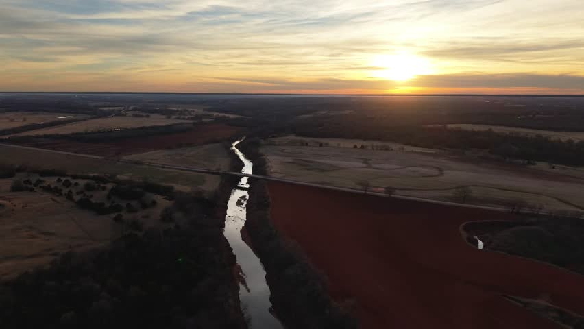Aerial drone view of winding river reflecting sunset light through rural farmland and fields in Oklahoma countryside landscape.