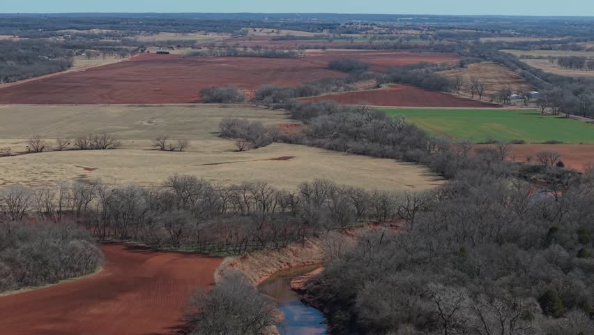 Aerial drone view of patchwork farmland and winding river cutting through winter fields and trees in rural Oklahoma landscape.
