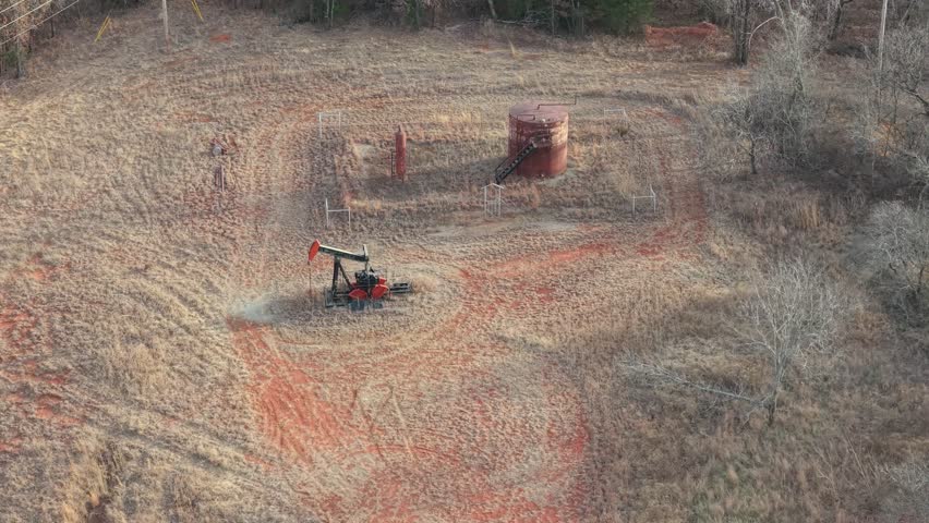 Aerial drone view of oil pumpjack operating beside storage tank in rural Oklahoma winter oilfield landscape.