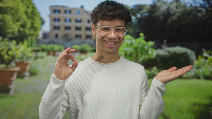 Young hispanic man points finger to palm in front of building in a sunlit green park; warm invitation.